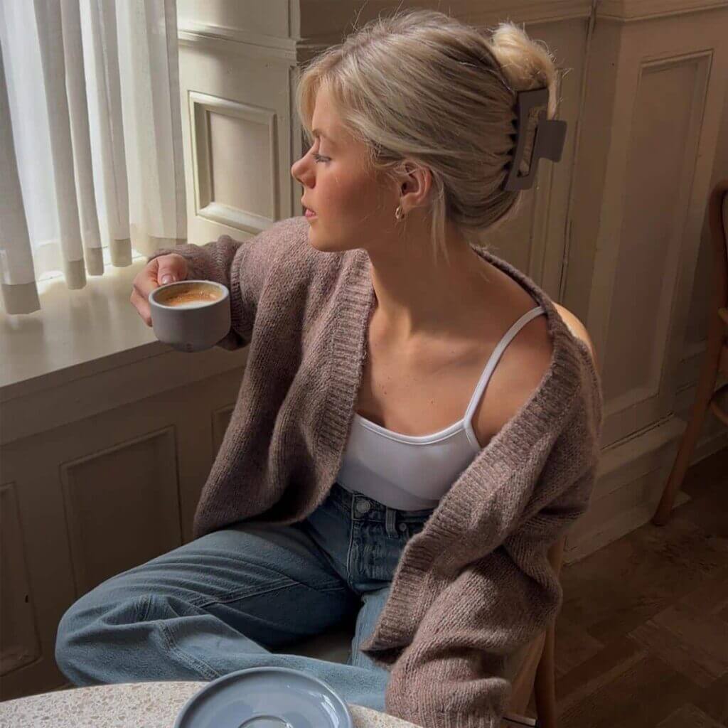 Ljusgrå hårklämma i knut – stylad i blont hår på modella i brun kofta, dricker kaffe vid fönster